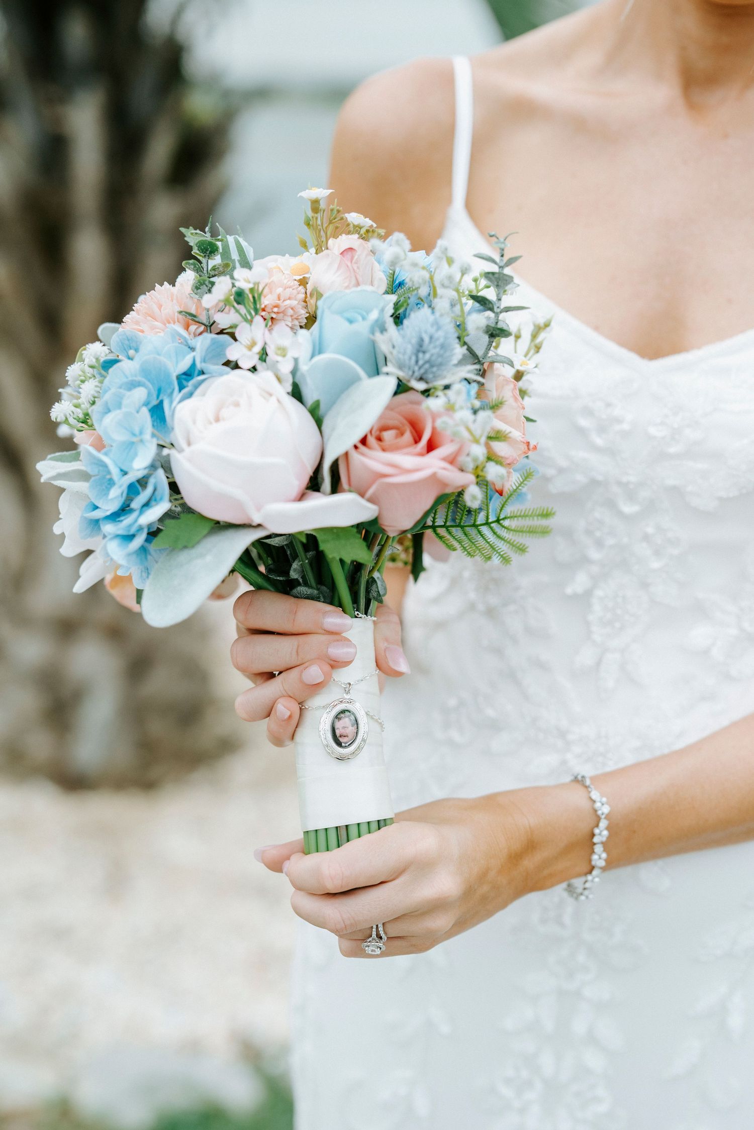 Close-up of pastel blue and pink wedding bouquet with vintage brooch detail against white lace dress.