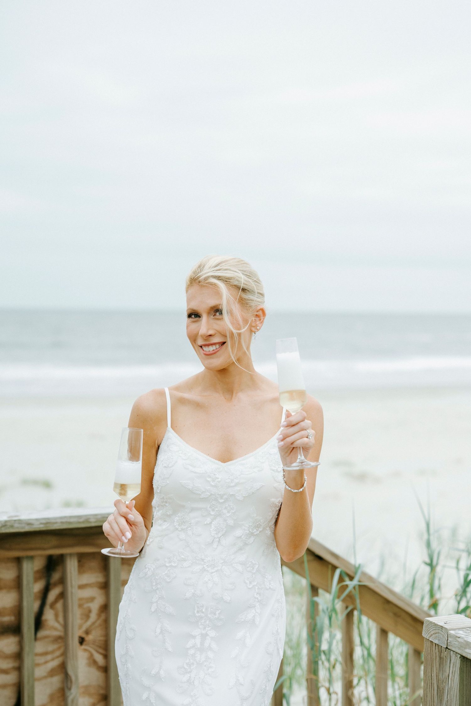 Bride in white lace dress smiles while holding champagne glass on coastal boardwalk.