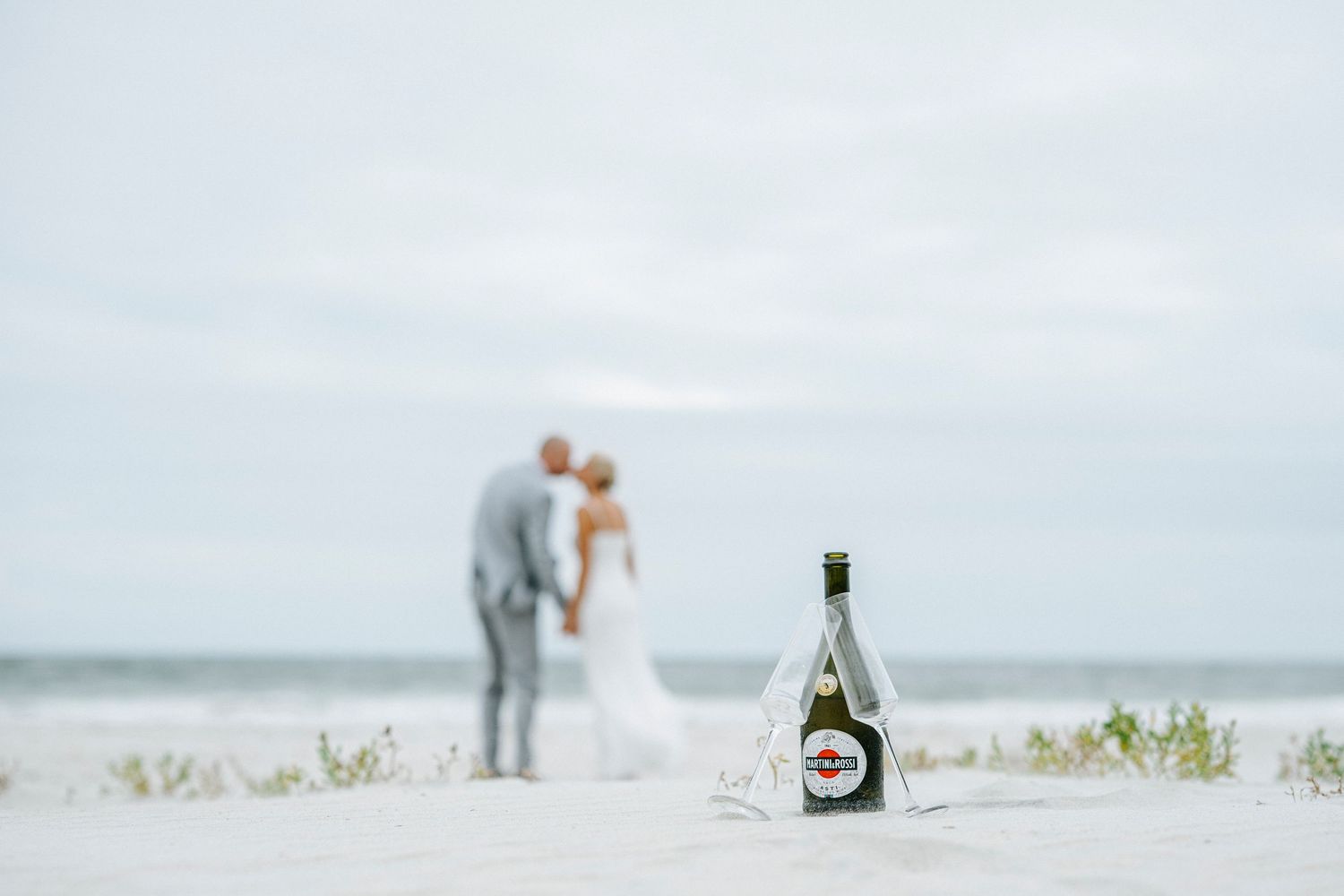 Newlywed couple walking together on a misty beach with champagne and a floral bouquet.