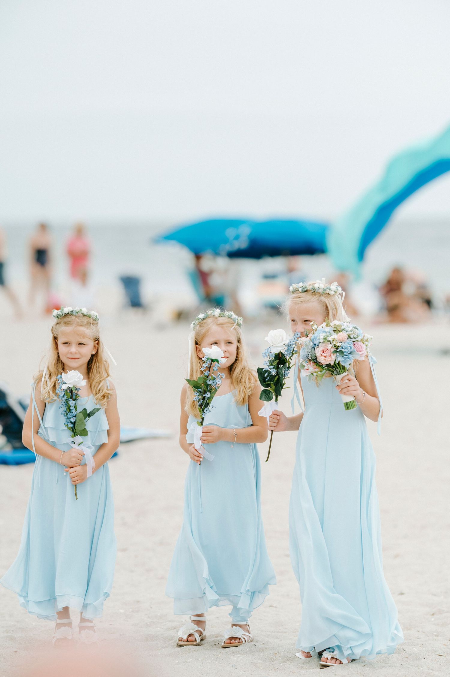 Three flower girls in flowing light blue dresses holding white floral bouquets stand on a sunny beach.