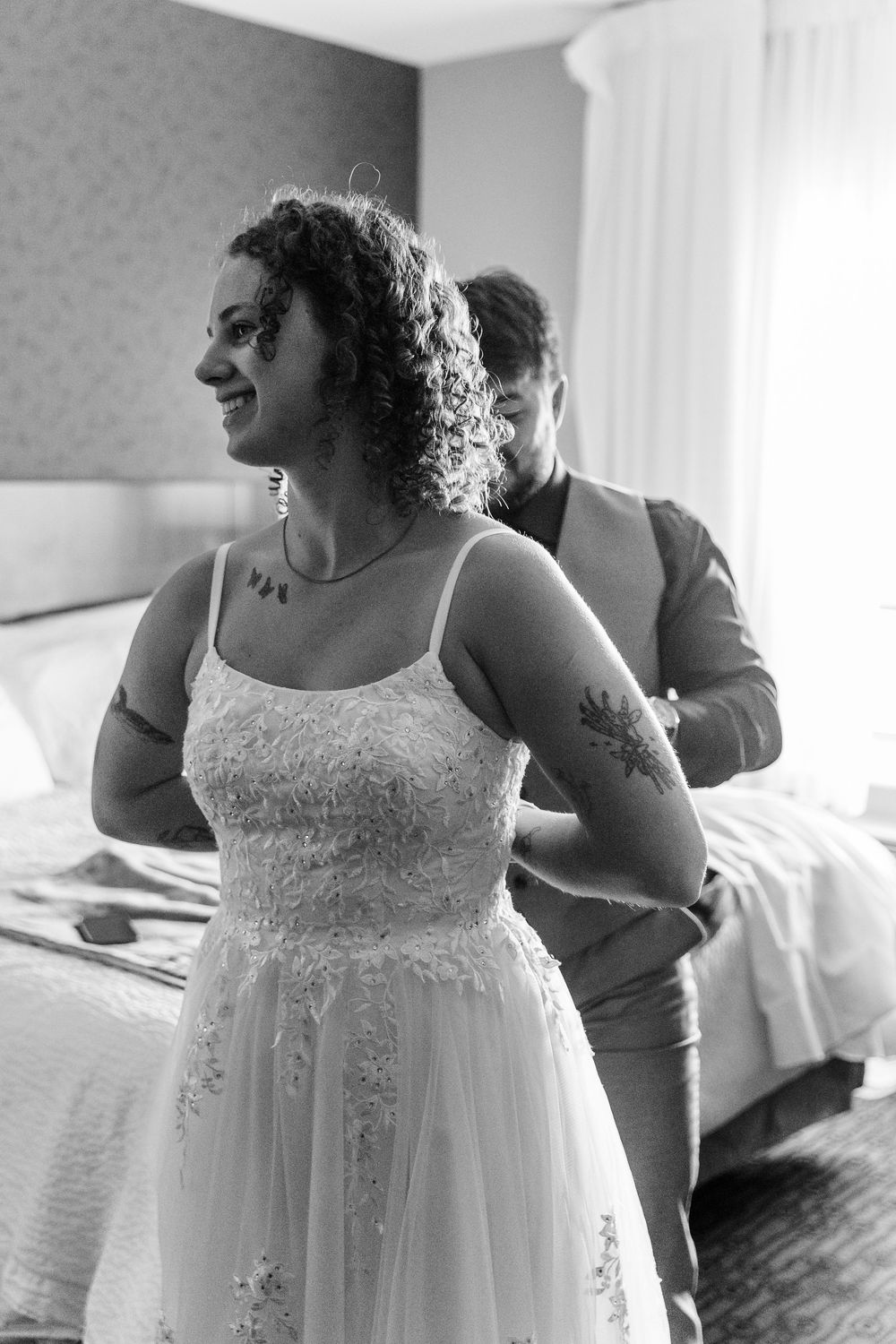 A person in a white lace wedding dress stands in a room with natural light filtering through the window.