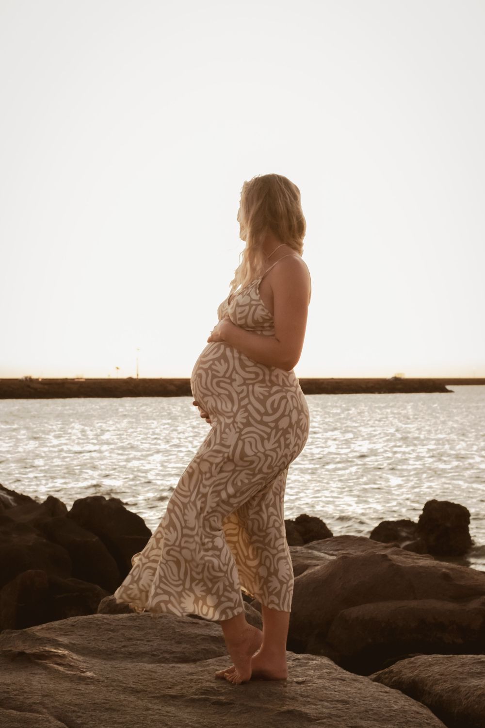 Expectant mother in floral dress posing on coastal rocks during golden hour.