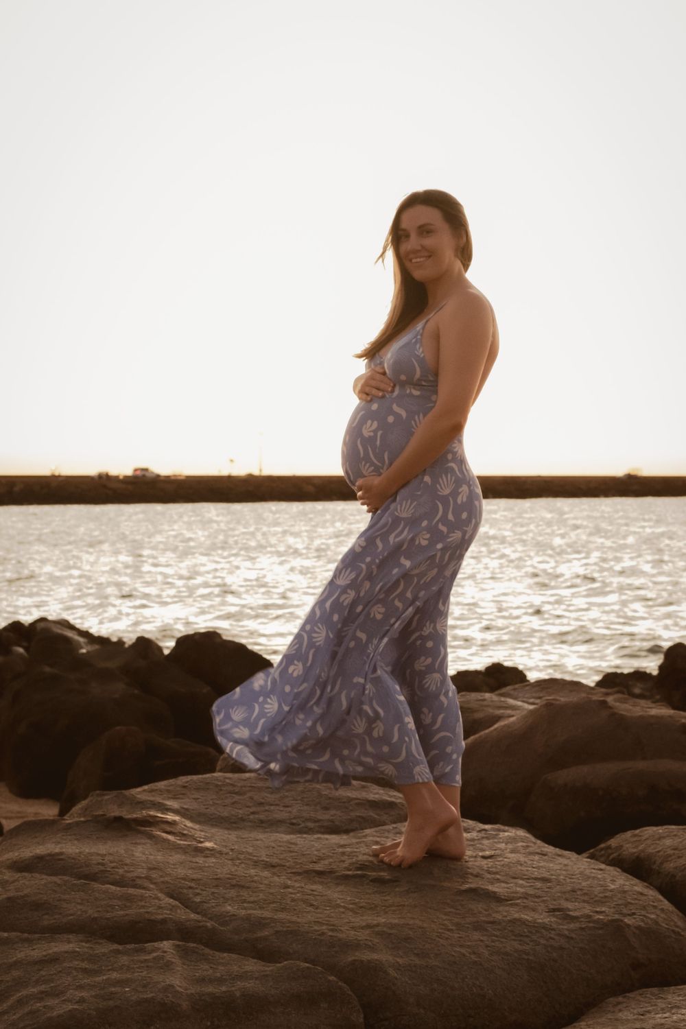 A maternity photoshoot on rocky beach at sunset featuring a flowing dress.