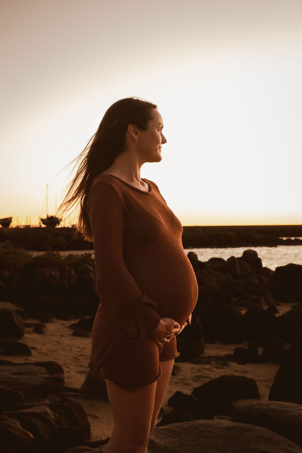 Sunset maternity portrait with figure in silhouette against ocean and rocks.