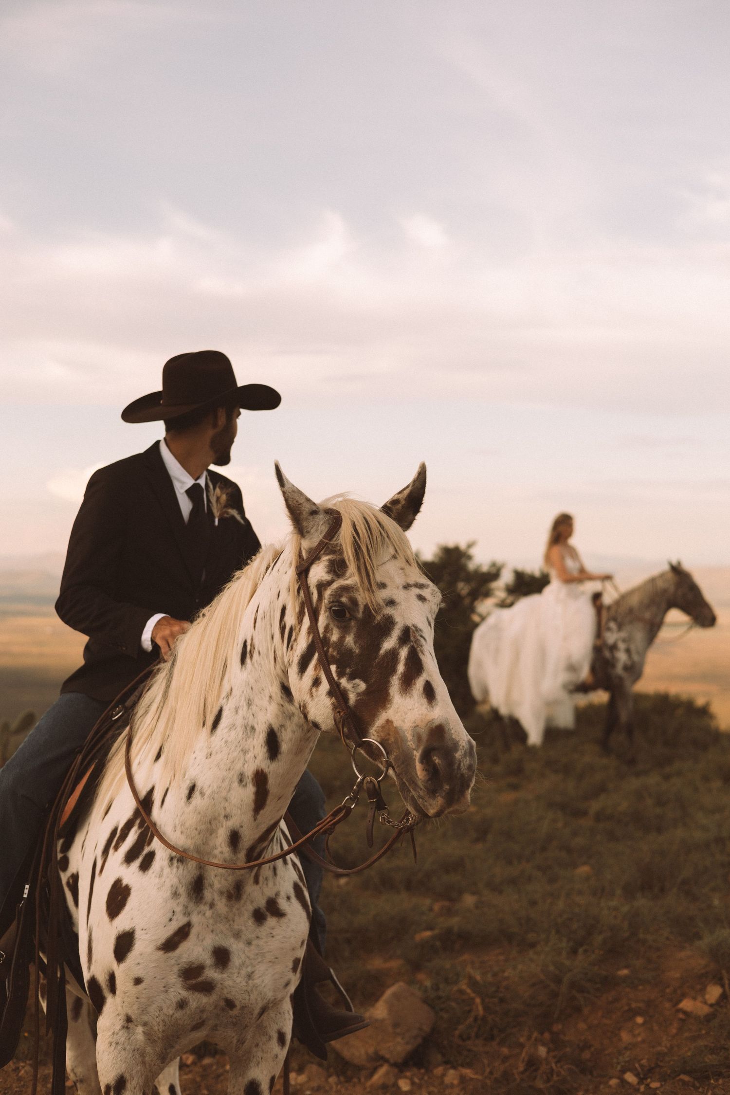 A cowboy on horseback rides a spotted white horse across rugged terrain at sunset.