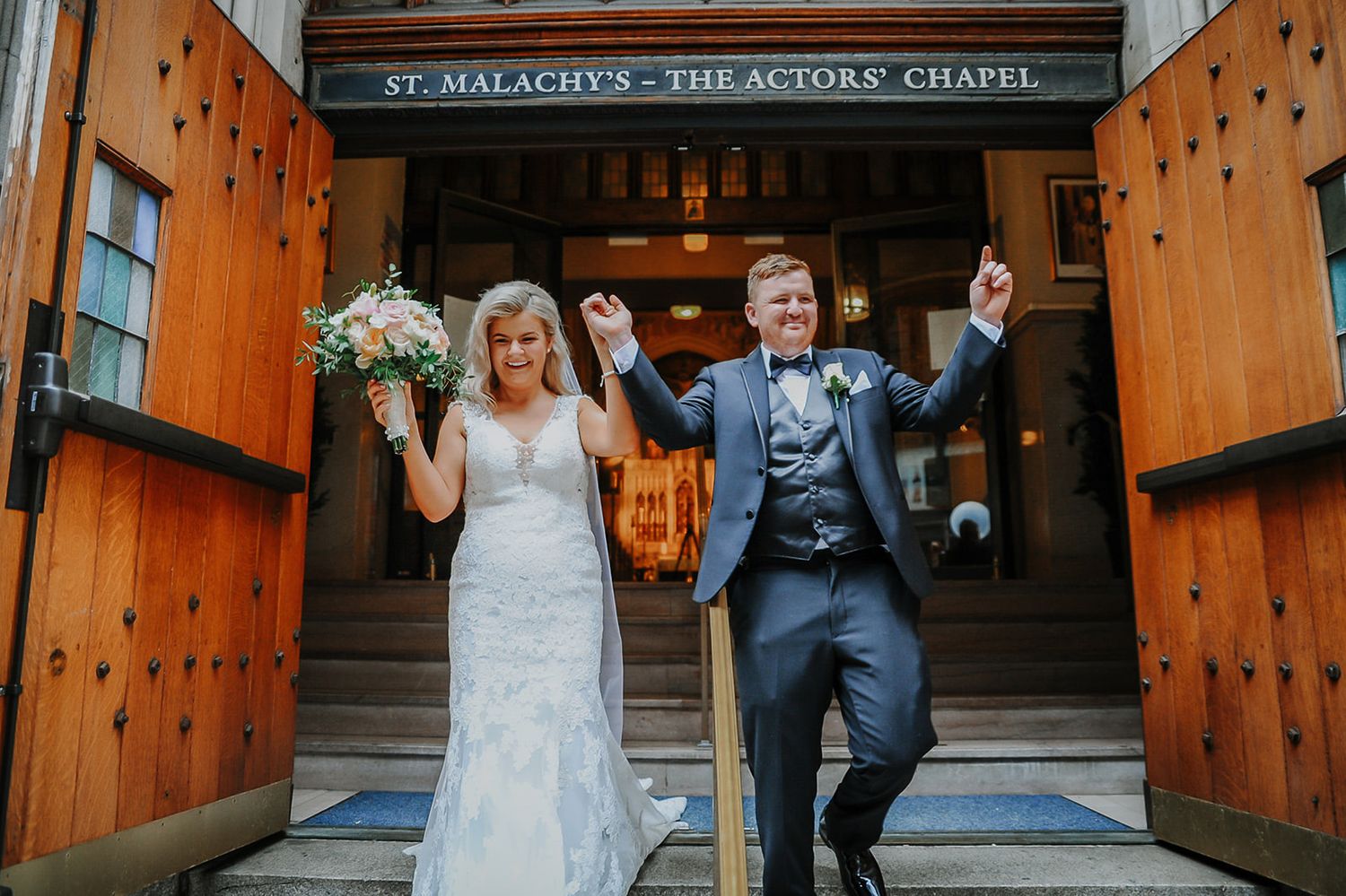 Joyful couple celebrating their wedding day outside St. Malachy's - The Actors' Chapel with raised arms.