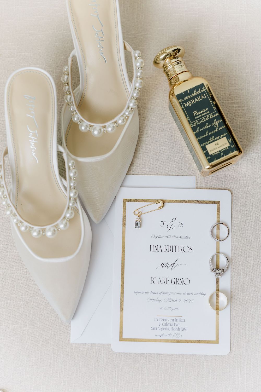 Wedding details flatlay with white shoes, invitation suite, and rings on beige background.