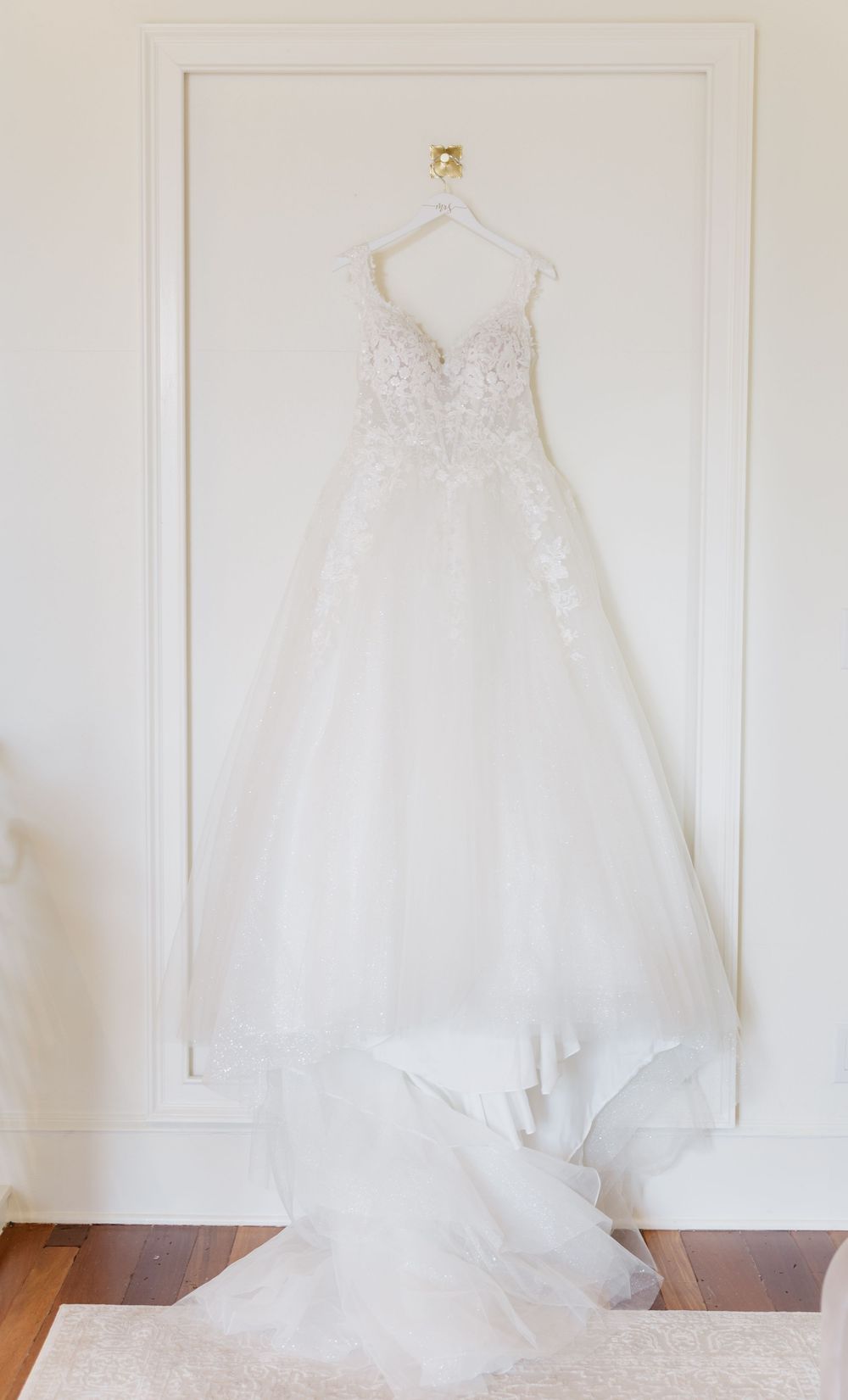 An elegant white wedding dress with lace details hangs on display against a white wall.