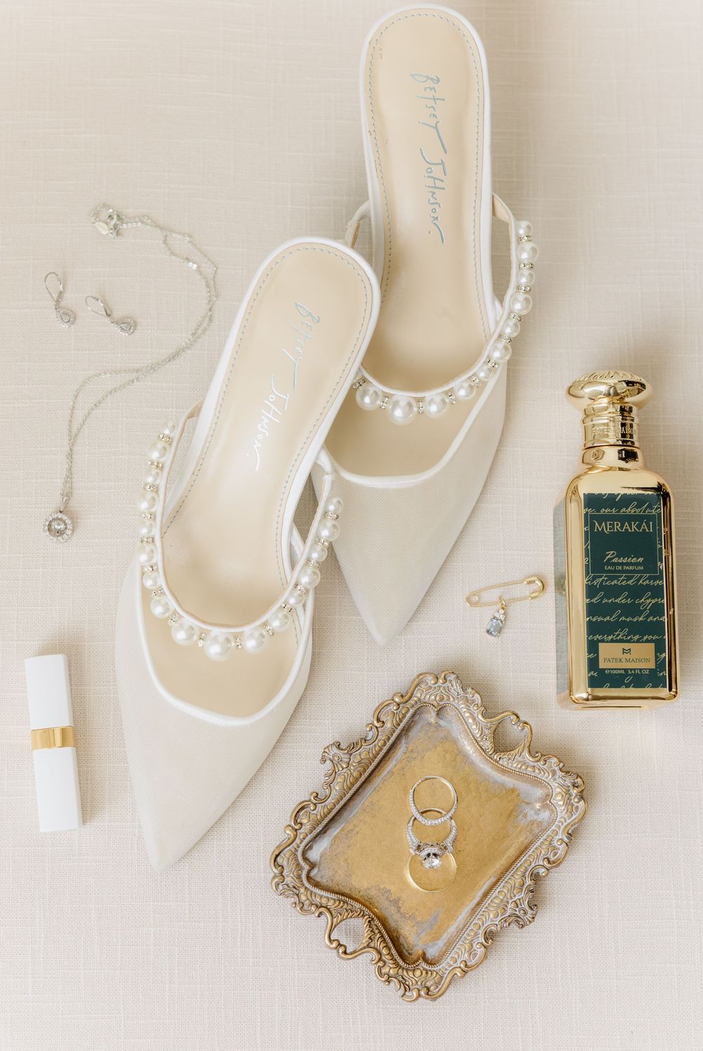 Wedding accessories flatlay featuring white pearl-embellished slippers, perfume bottle, and jewelry on ornate tray.