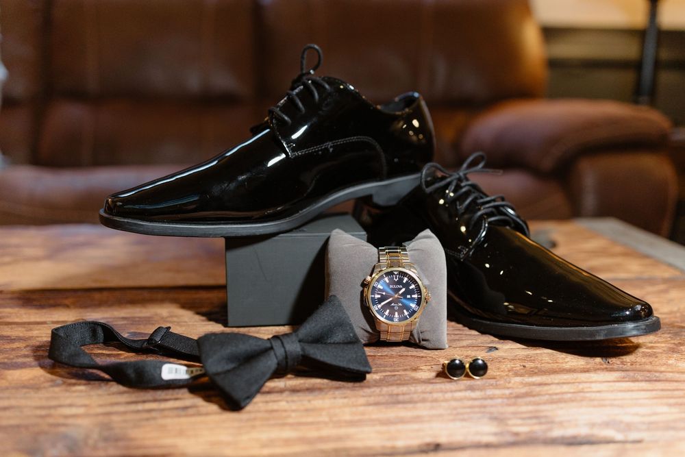 Black formal groom's accessories including dress shoes, bow tie and watch on wooden surface.