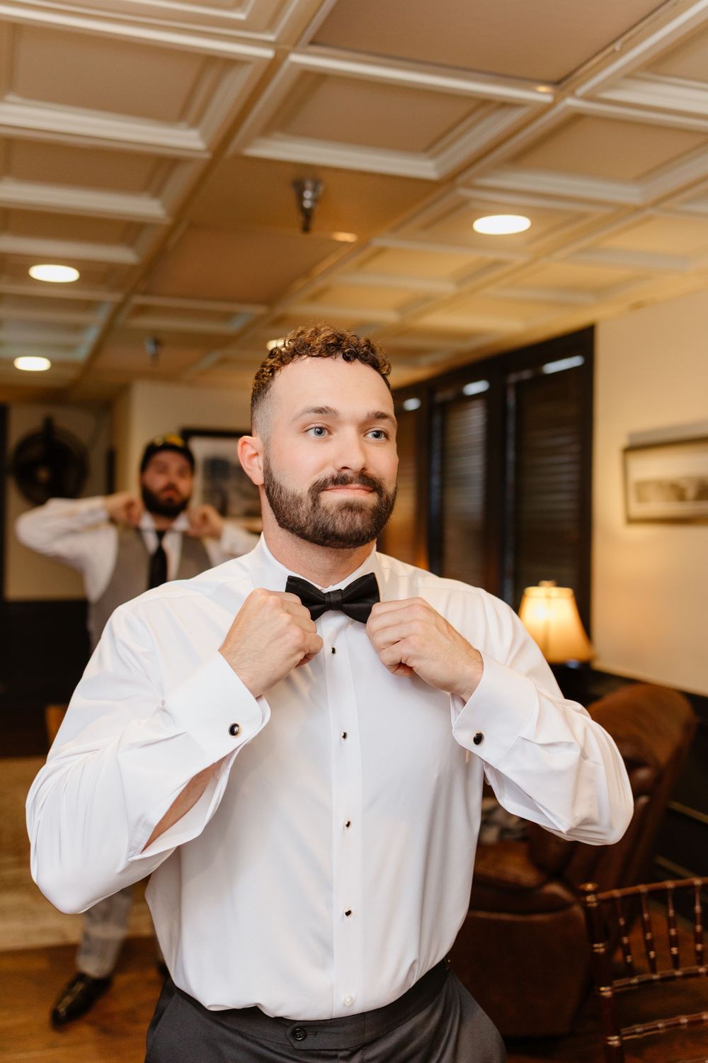 A groom adjusts his collar while getting ready in a warm-lit room with coffered ceiling.