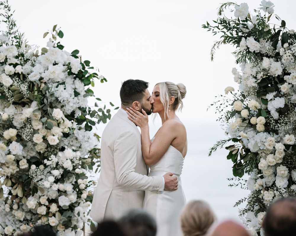Beautiful newlyweds share their first kiss at an elegant white floral decorated outdoor wedding ceremony.