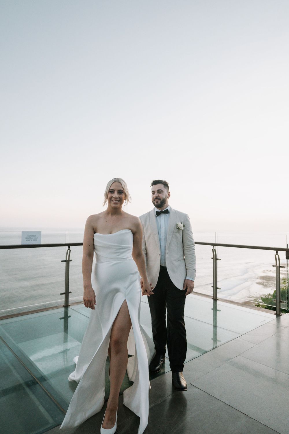 A couple poses for wedding photos on a modern balcony overlooking the ocean during sunset.