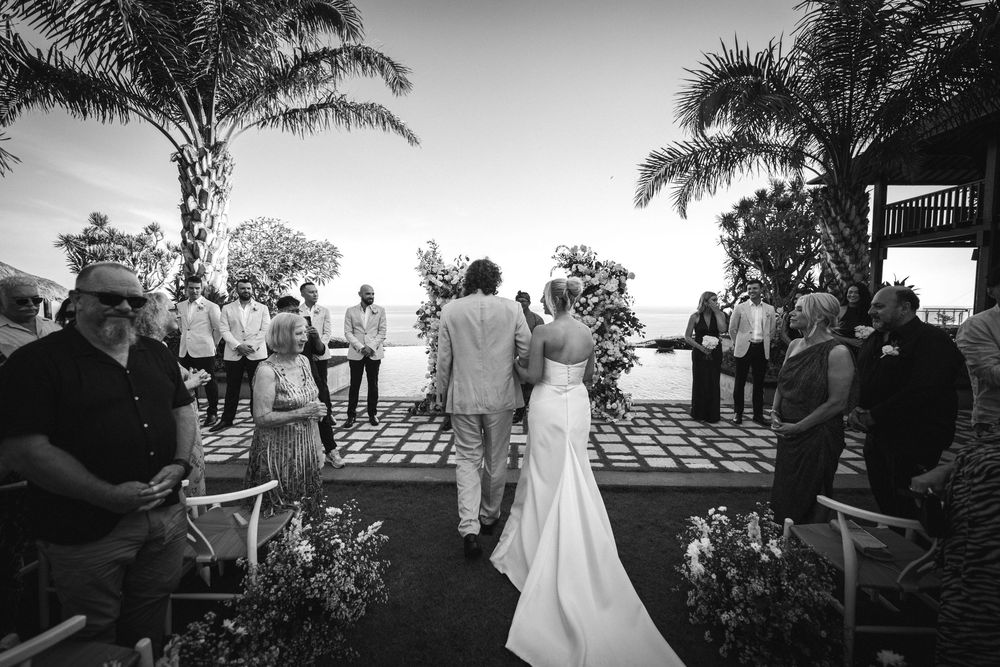 Black and white wedding ceremony photos show palm trees and guests gathered oceanside in a romantic outdoor setting.