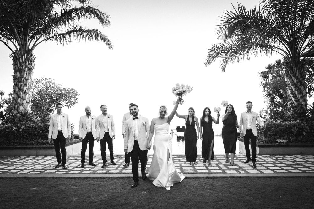 A black and white wedding party stands in a line on a tropical beach with palm trees silhouetted against the sky.