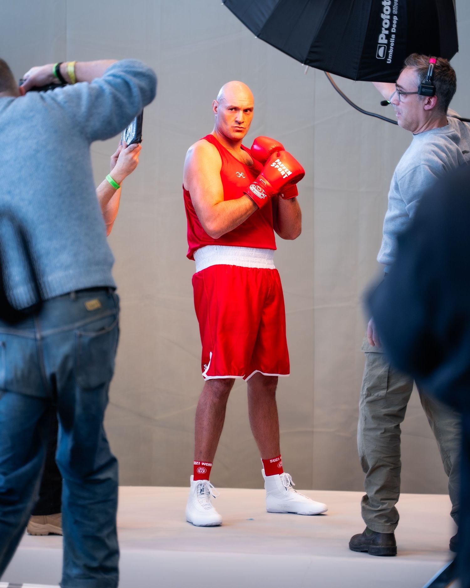 A person in red boxing attire and white shoes poses during a photo shoot against a gray backdrop.