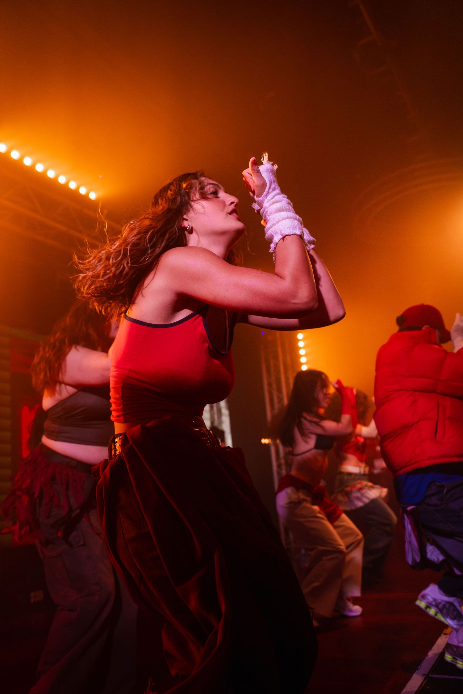 Performer in a red top dances energetically under warm orange stage lighting at a concert.