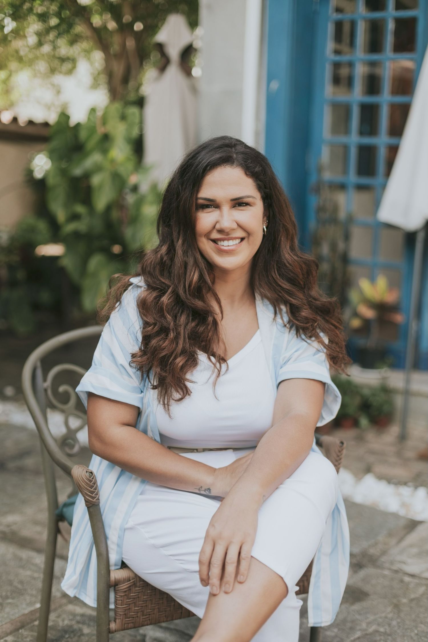 Portrait of a woman in white clothing sitting on a patio chair with blue doors and greenery in background on Maui, Hawaii.