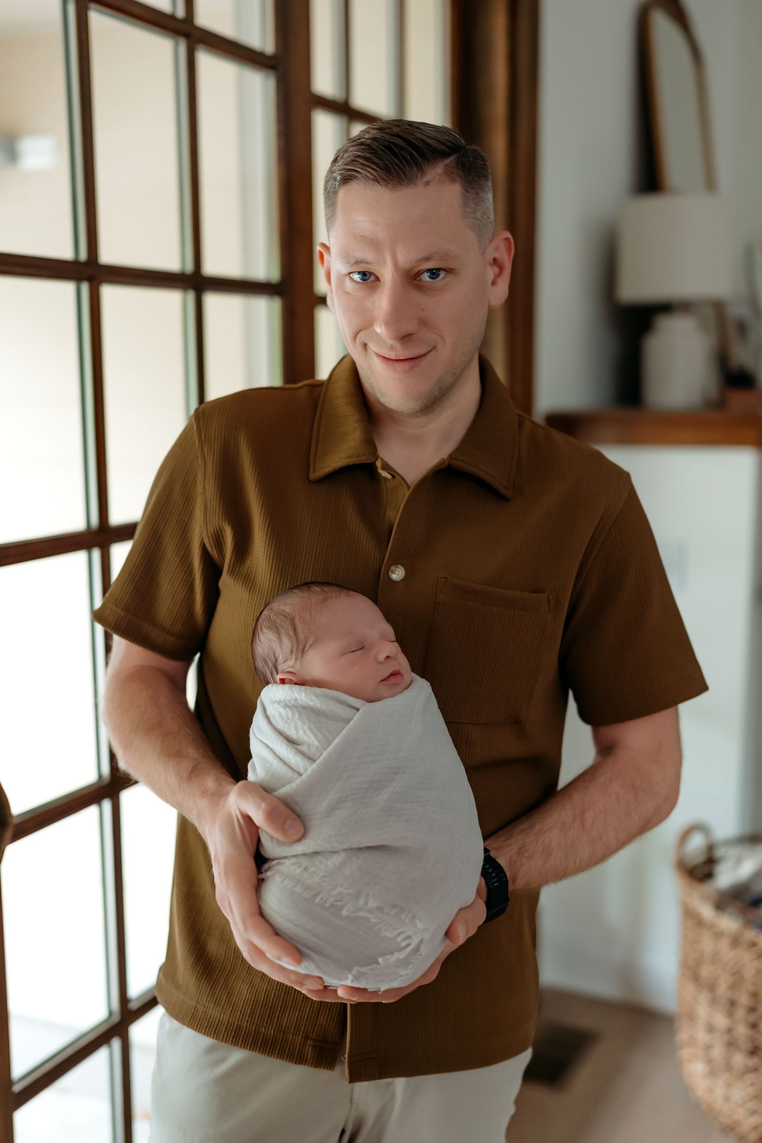 A person in a brown polo shirt holds a swaddled newborn baby while standing near windows in a bright room.