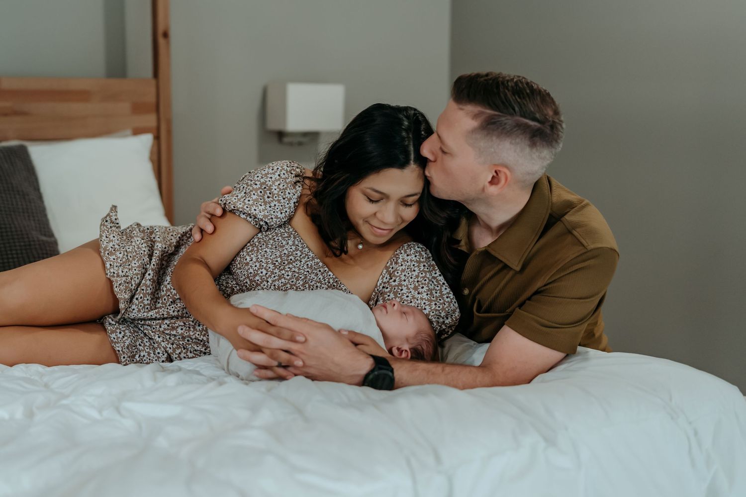 A couple shares an intimate moment while laying on a bed with their newborn baby between them.