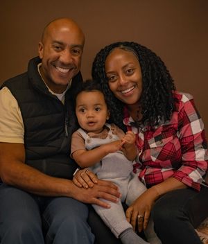 A family of three sits close together on a brown couch sharing warm smiles and embraces.