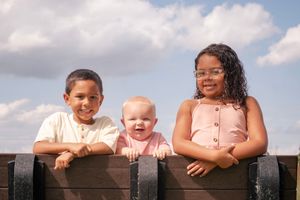 Children leaning over a wooden fence in a sunny outdoor setting with clouds in the sky.