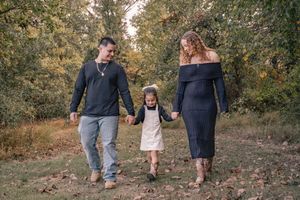 A family walks hand in hand through a wooded area on an autumn day wearing casual attire.