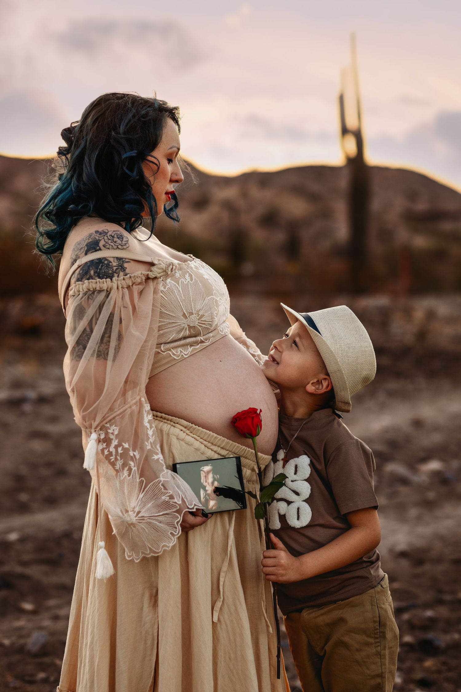 A touching maternity photo shows pregnant figure in flowing dress with child holding red flower in desert setting at sunset.