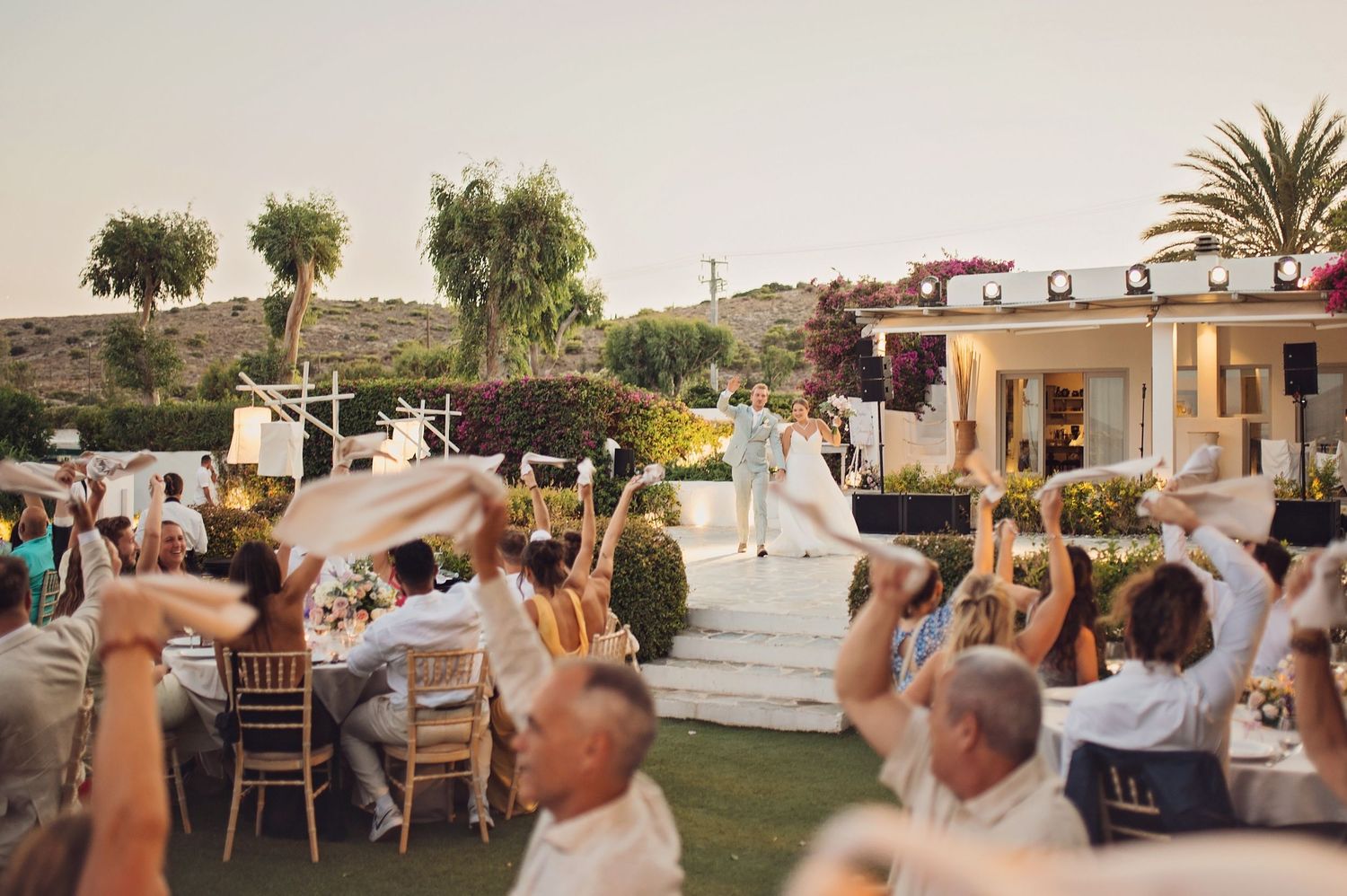 Wedding guests raise their arms in celebration during an outdoor reception at sunset.