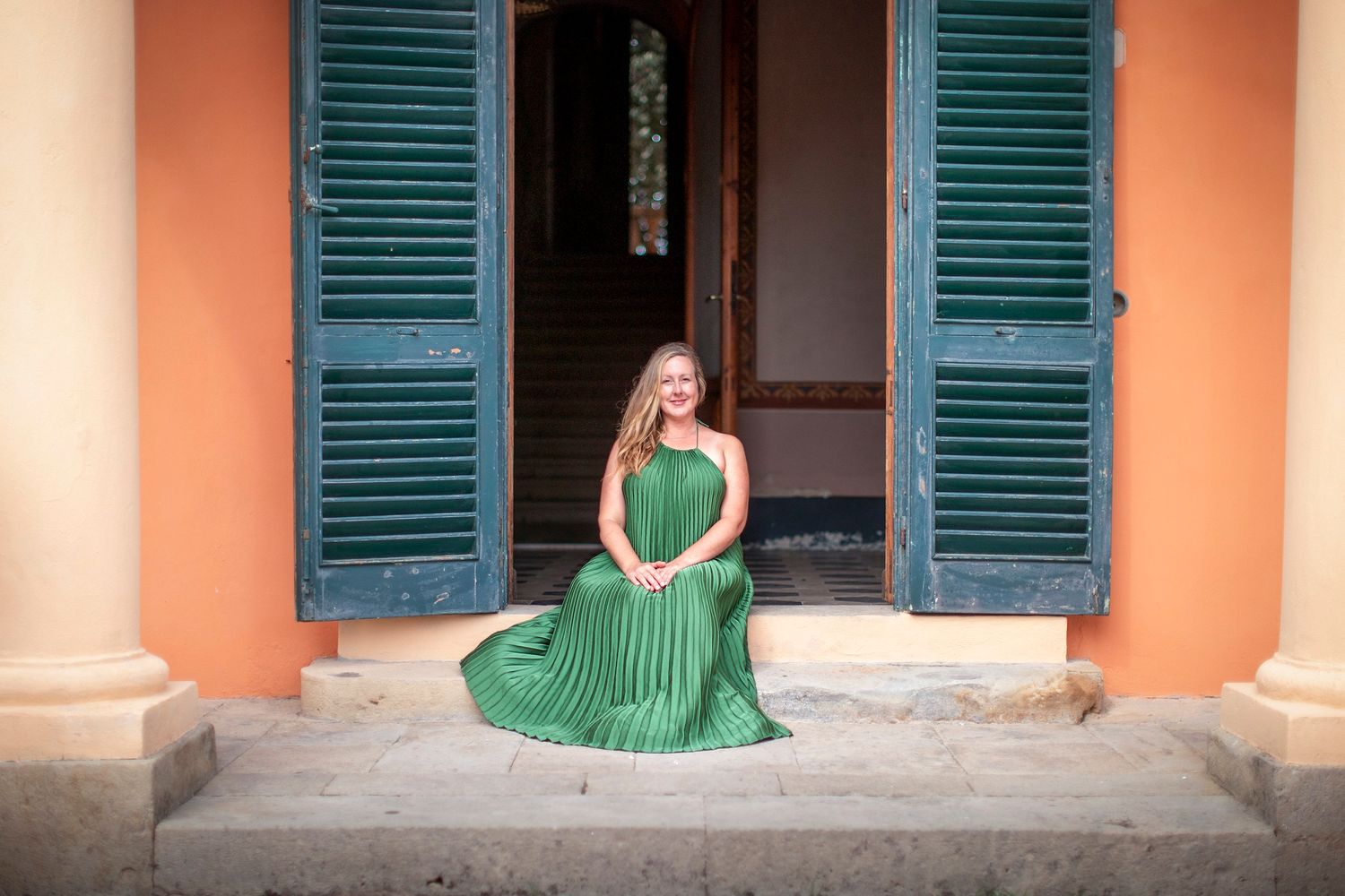 Woman in elegant green pleated dress sitting on steps between blue shutters of a colorful colonial building.