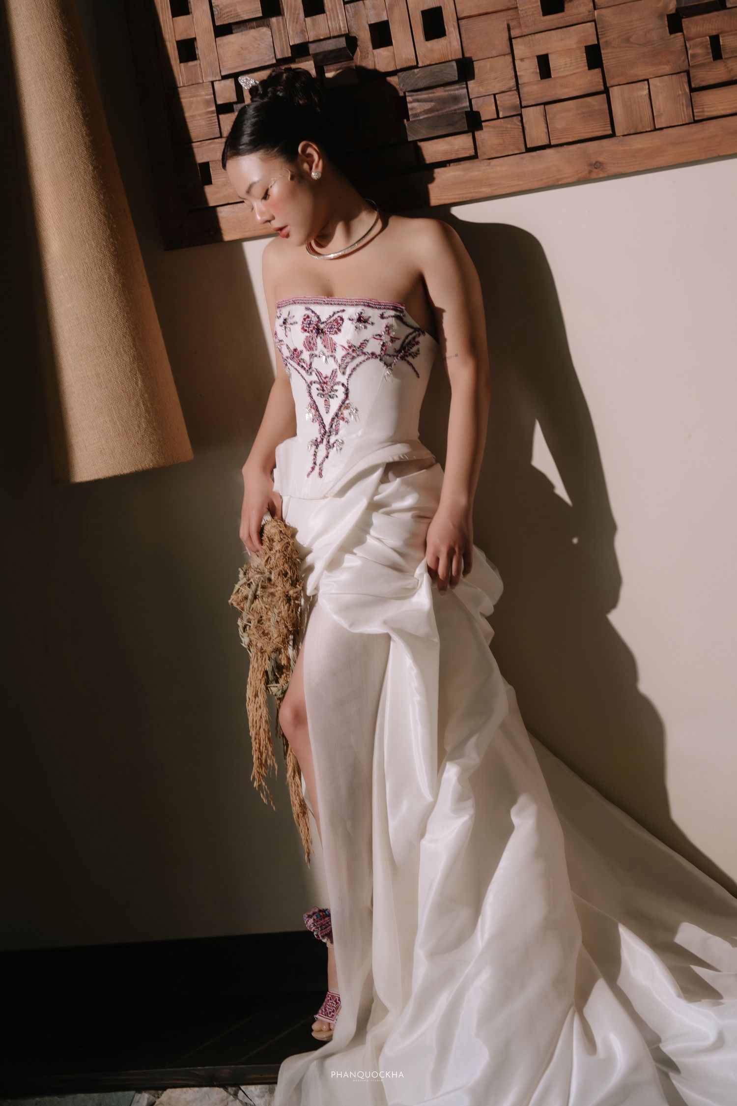 Bride in white strapless embroidered gown with thigh slit holding dried bouquet indoors.
