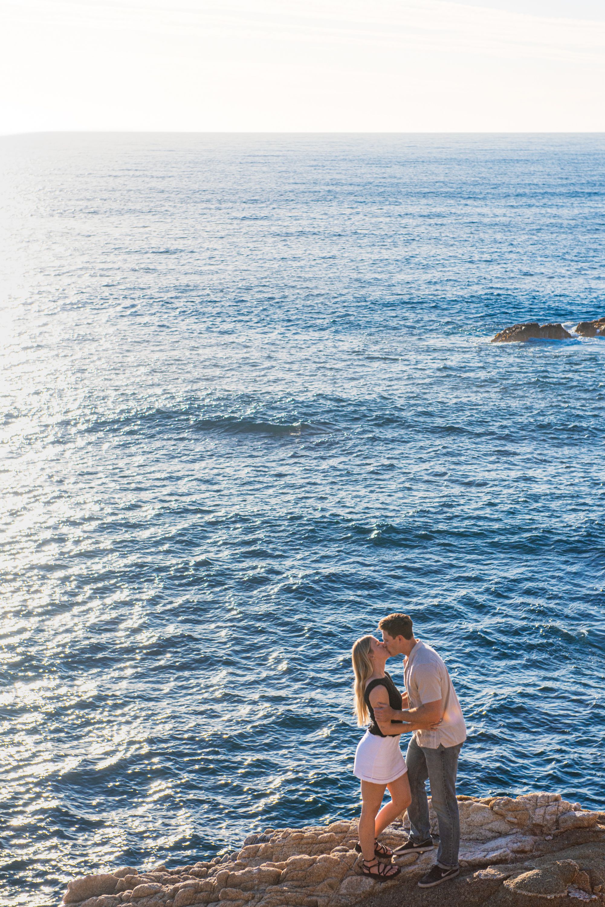 Romantic couple kissing on rocky coastal cliffs with stunning blue ocean waves in the background.