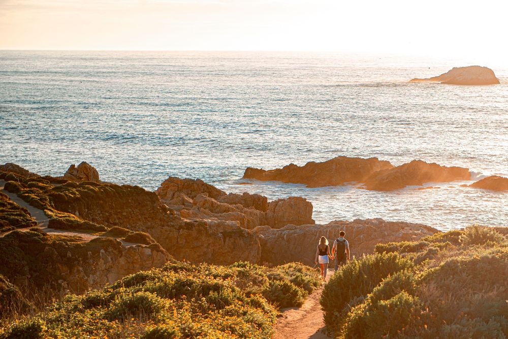 Two people walk along a rocky coastal path at golden hour with ocean views and distant rock formations.