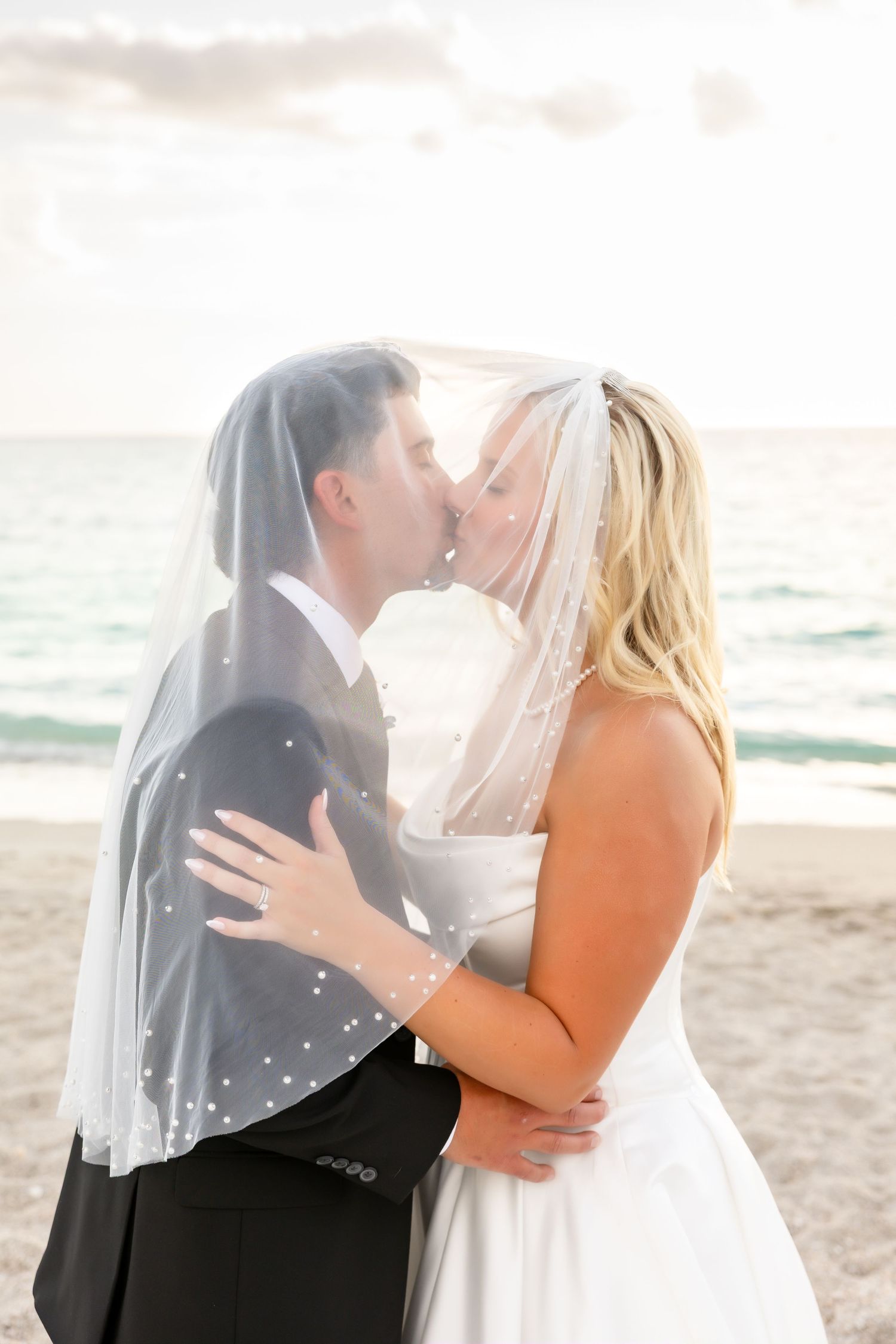 Bride and groom sharing a romantic kiss under a pearl-dotted wedding veil on a sunny beach.