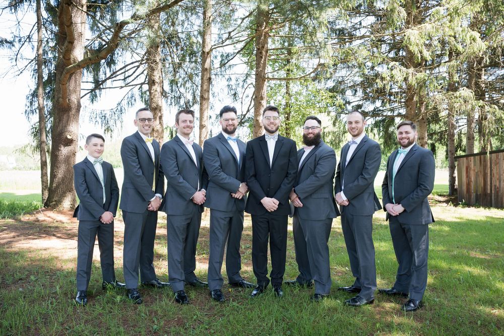A line of groomsmen in matching navy suits pose playfully amongst trees in a garden setting for wedding photos.