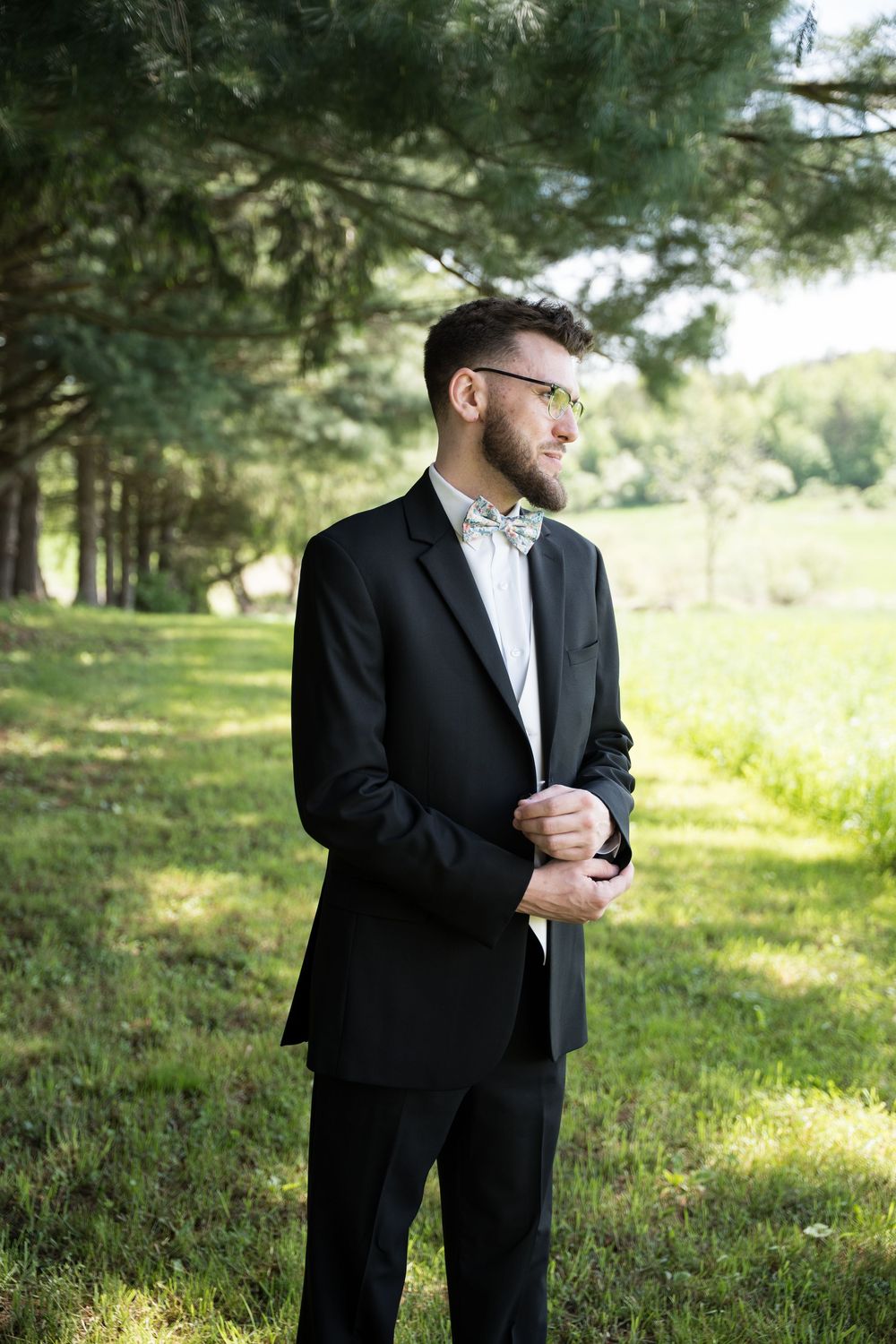 A figure in a black suit and glasses stands thoughtfully in a sunlit outdoor setting among pine trees.