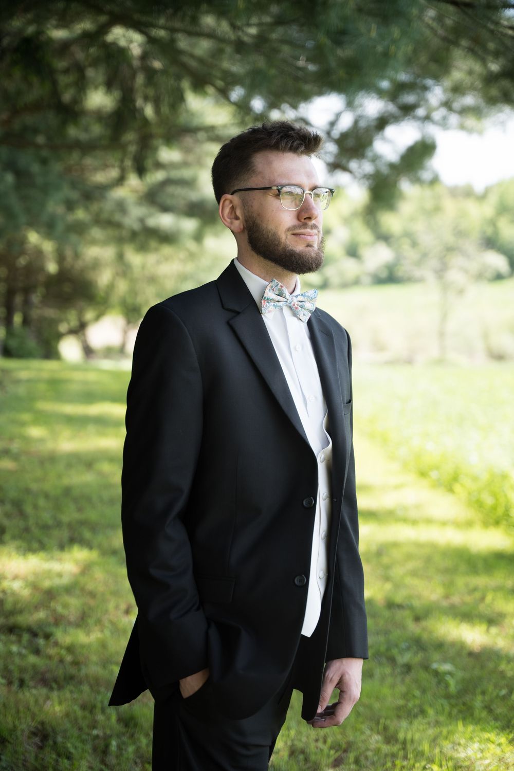 A groom in a black tuxedo and white bowtie poses outdoors in a sun-dappled garden setting.