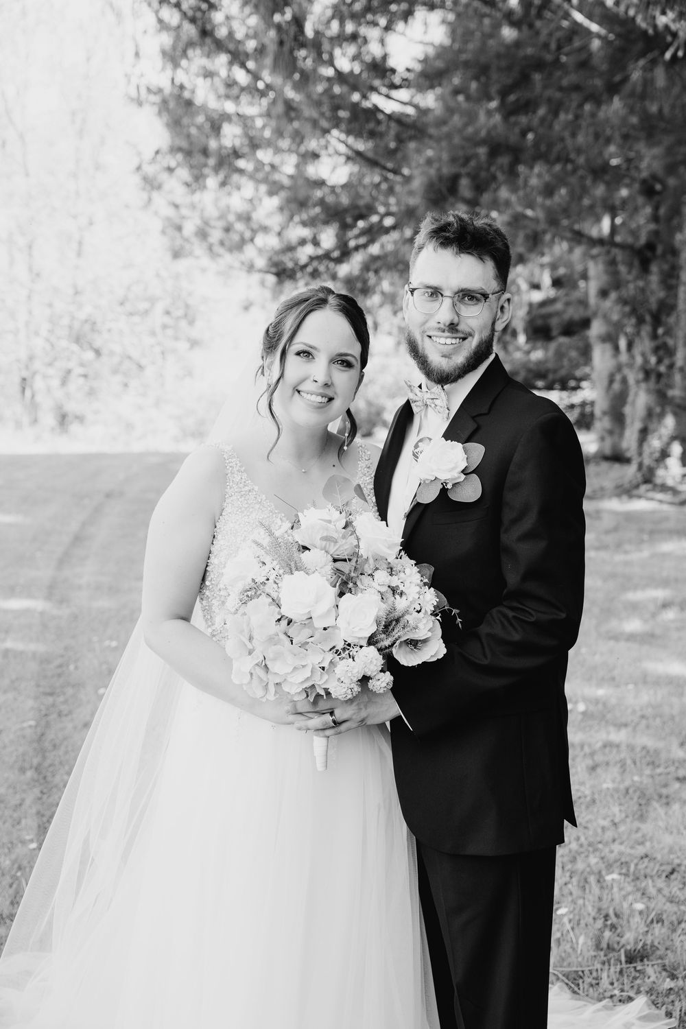 Newly married couple embraces outdoors with white bouquet and formal attire in natural setting.