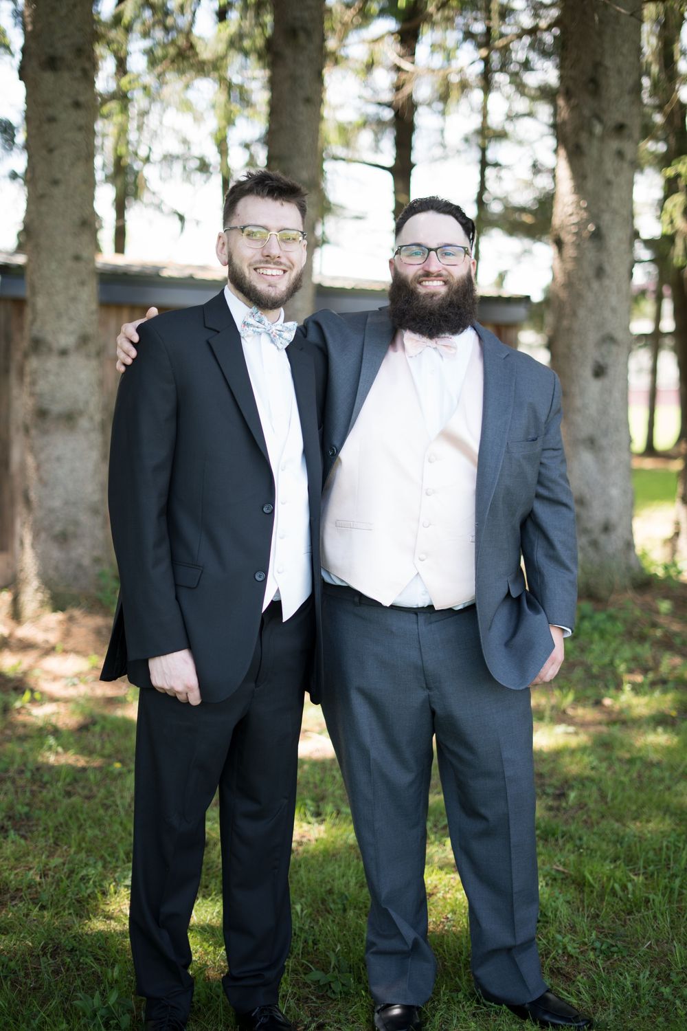 Two people in formal attire stand close together smiling outdoors in a wooded setting during daytime.