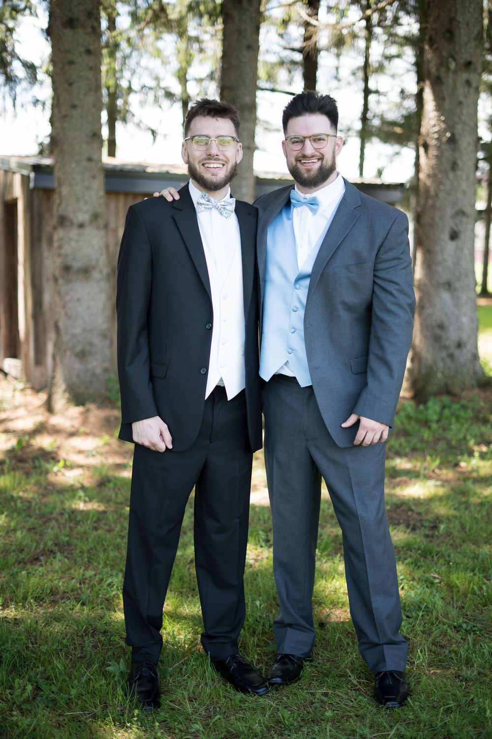 Wedding party members pose casually in tuxedos and suits among trees on grassy ground.