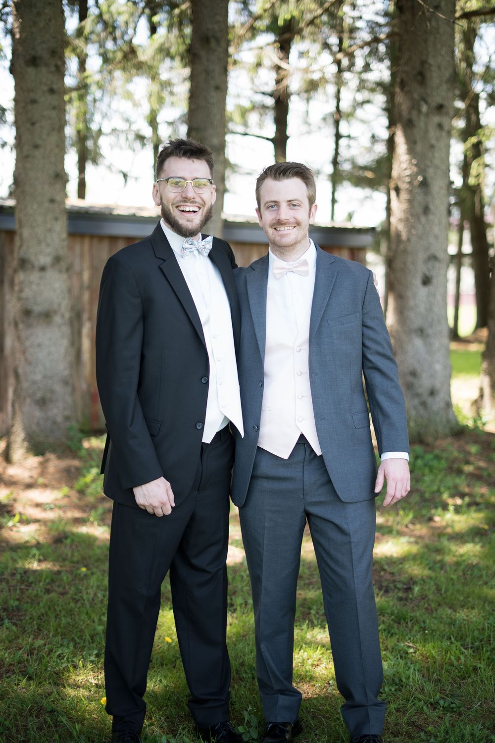 Group stands together in formal attire for wedding photos in a wooded outdoor setting.