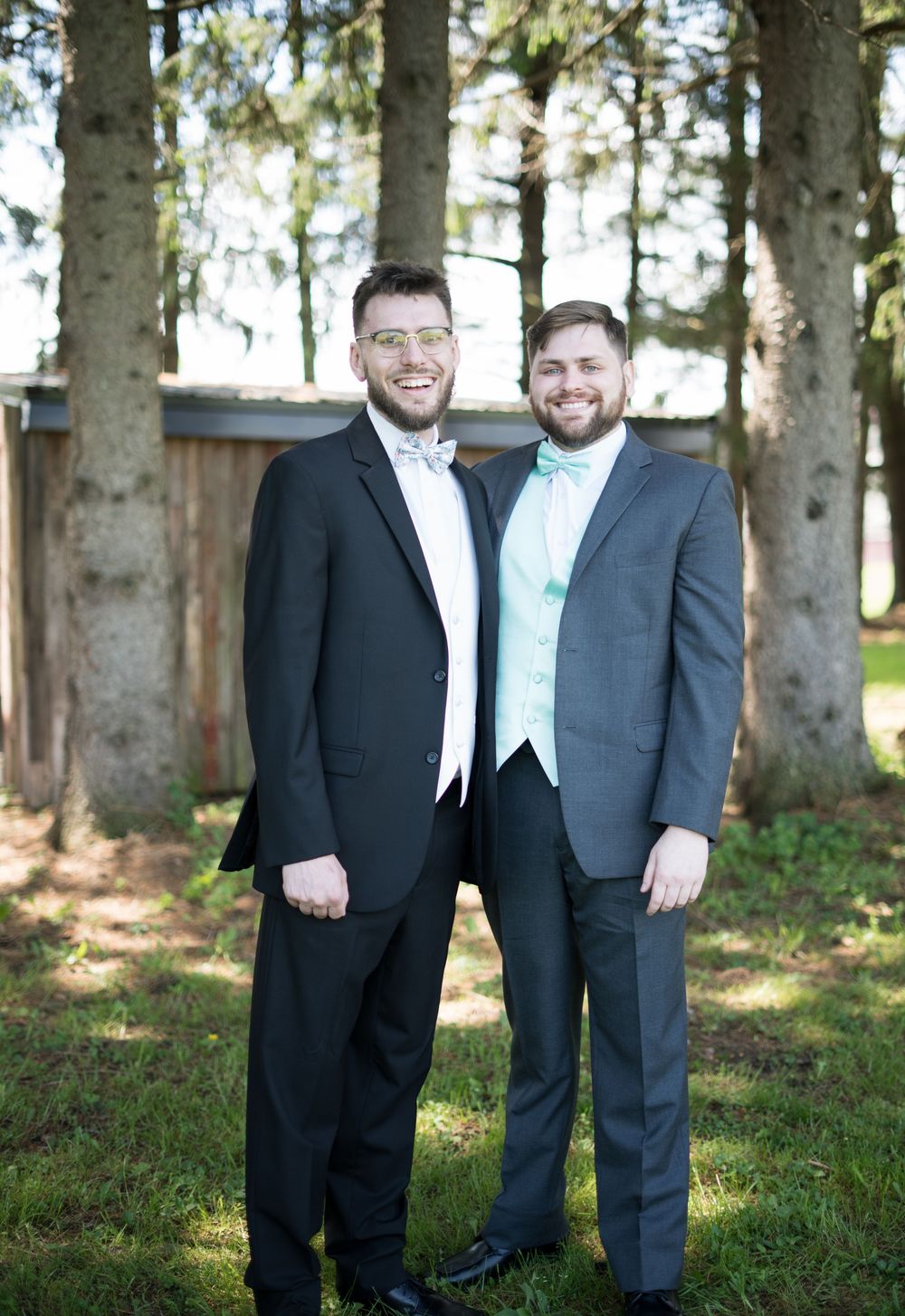 Two people in formal suits stand together outdoors in a wooded setting.