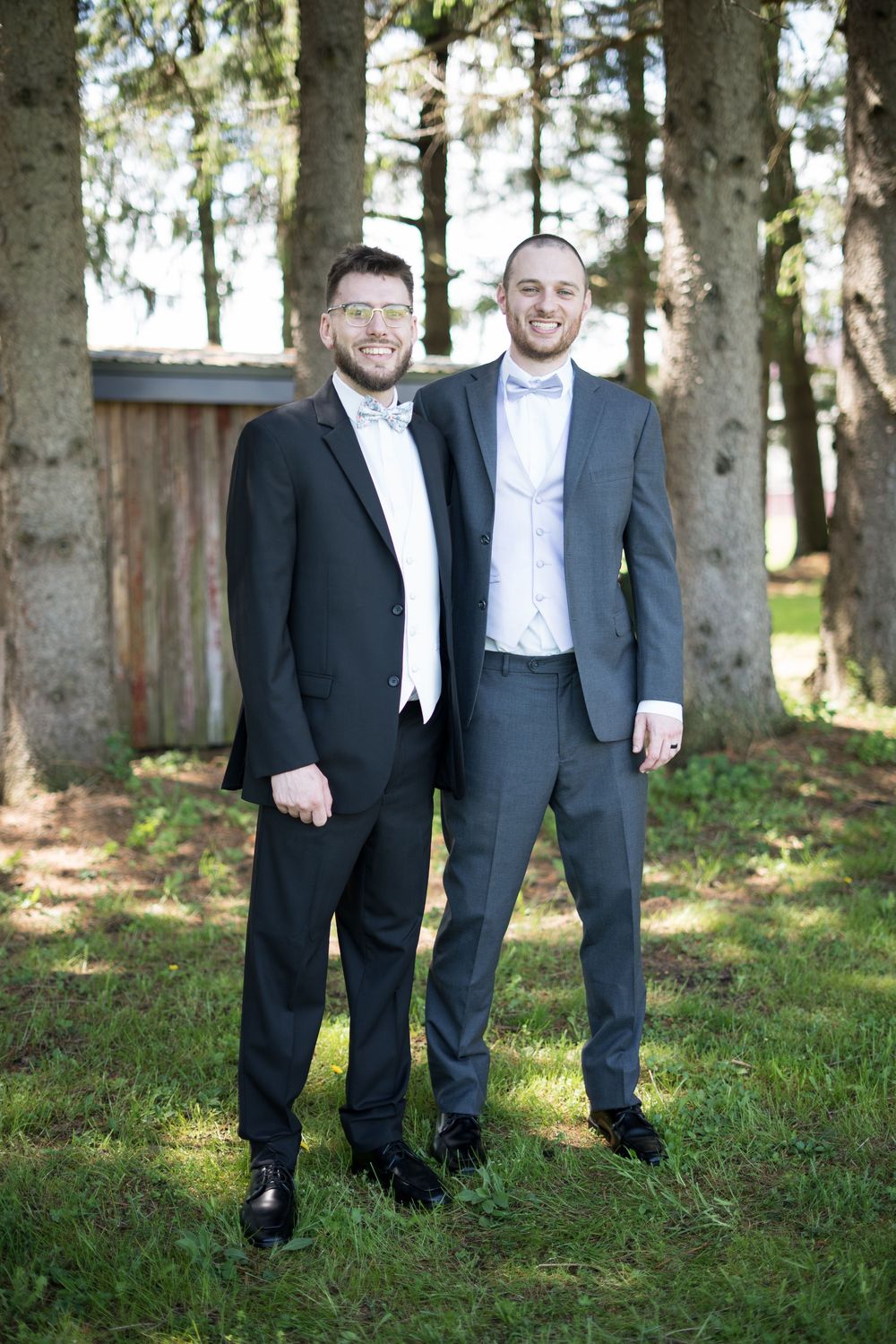 Two men in tuxedos standing together in a wooded outdoor setting with natural lighting and green grass.