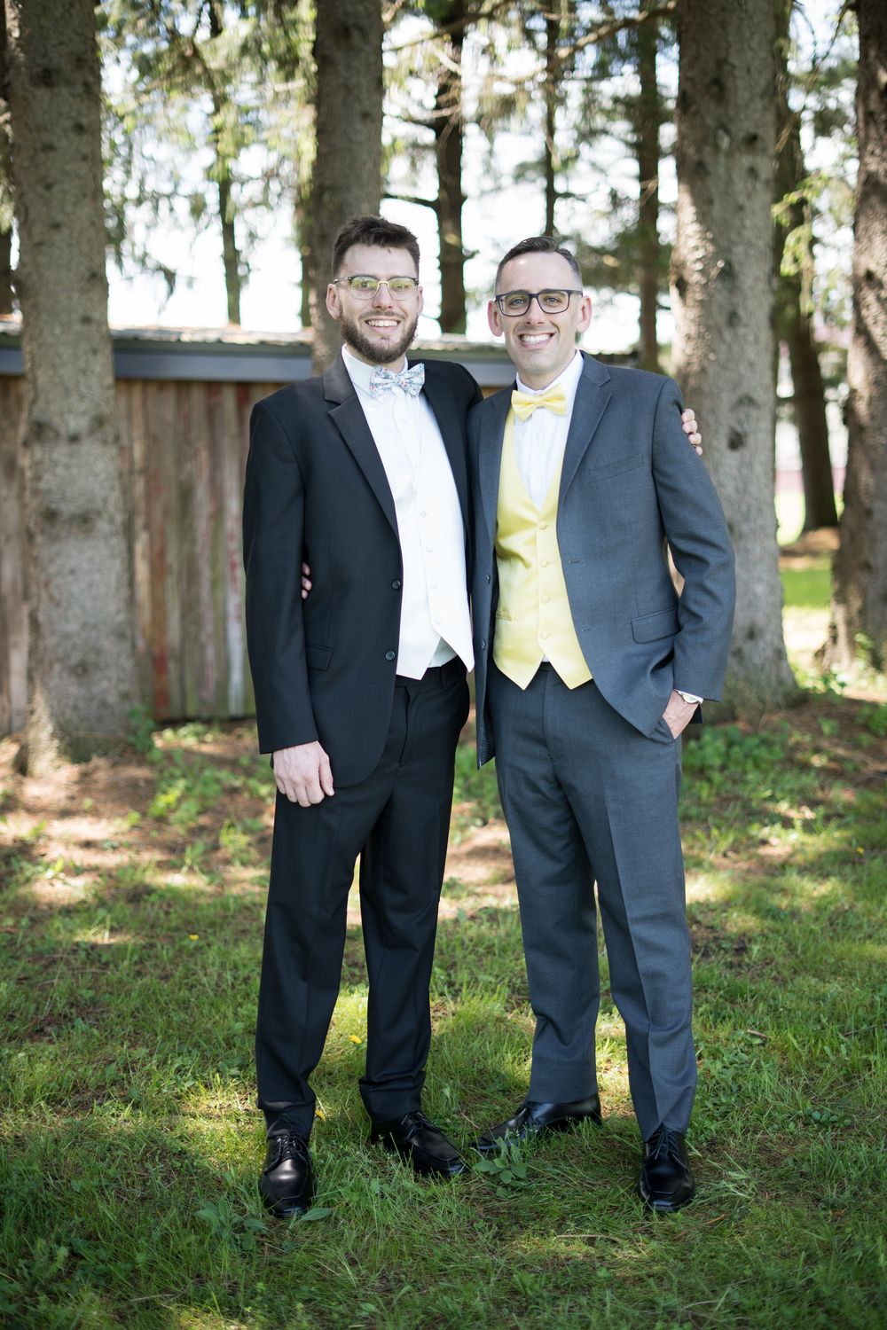 Two people in formal suits standing together outdoors on grass with trees in the background.