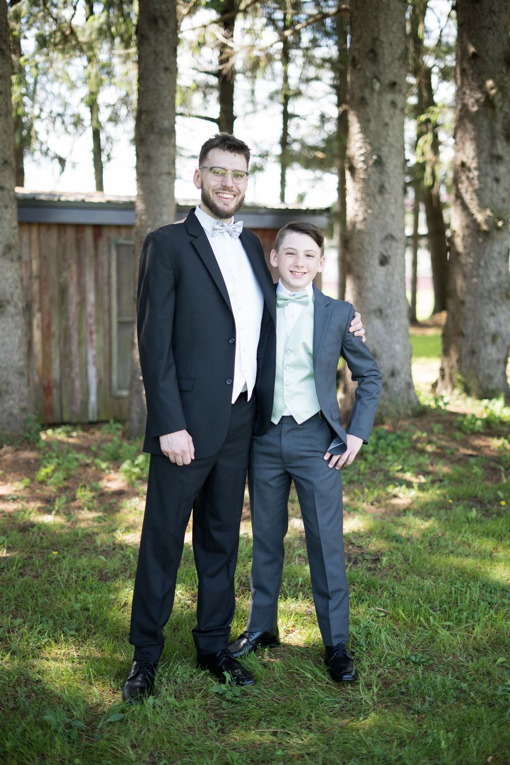 Two people wearing formal attire stand in front of tall trees with dappled sunlight filtering through the leaves.