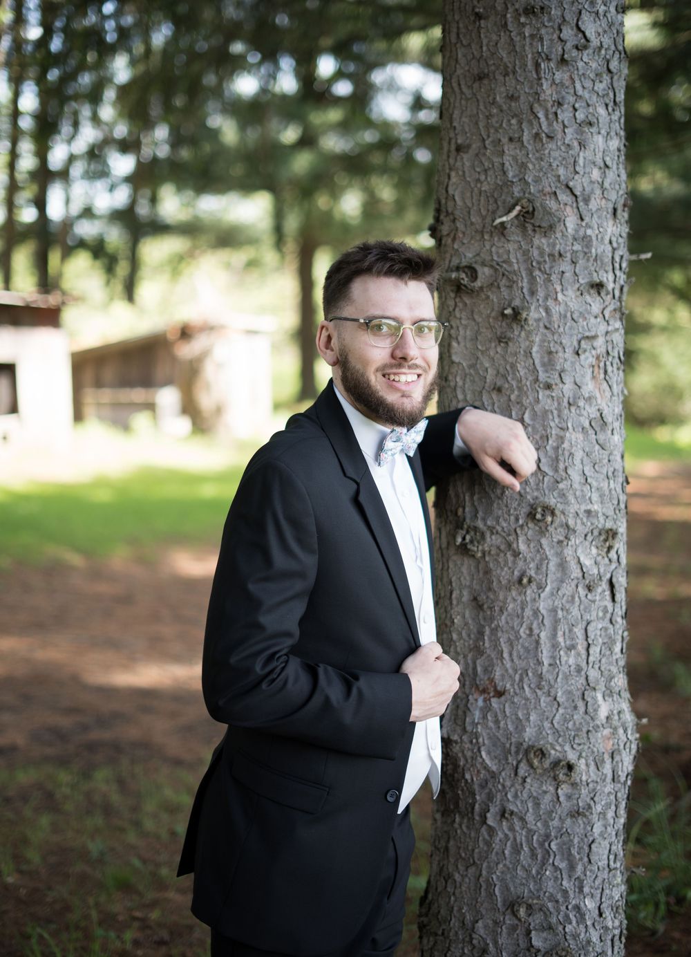 A groom in a classic black tuxedo and white bow tie poses playfully against a large tree trunk in a wooded setting.