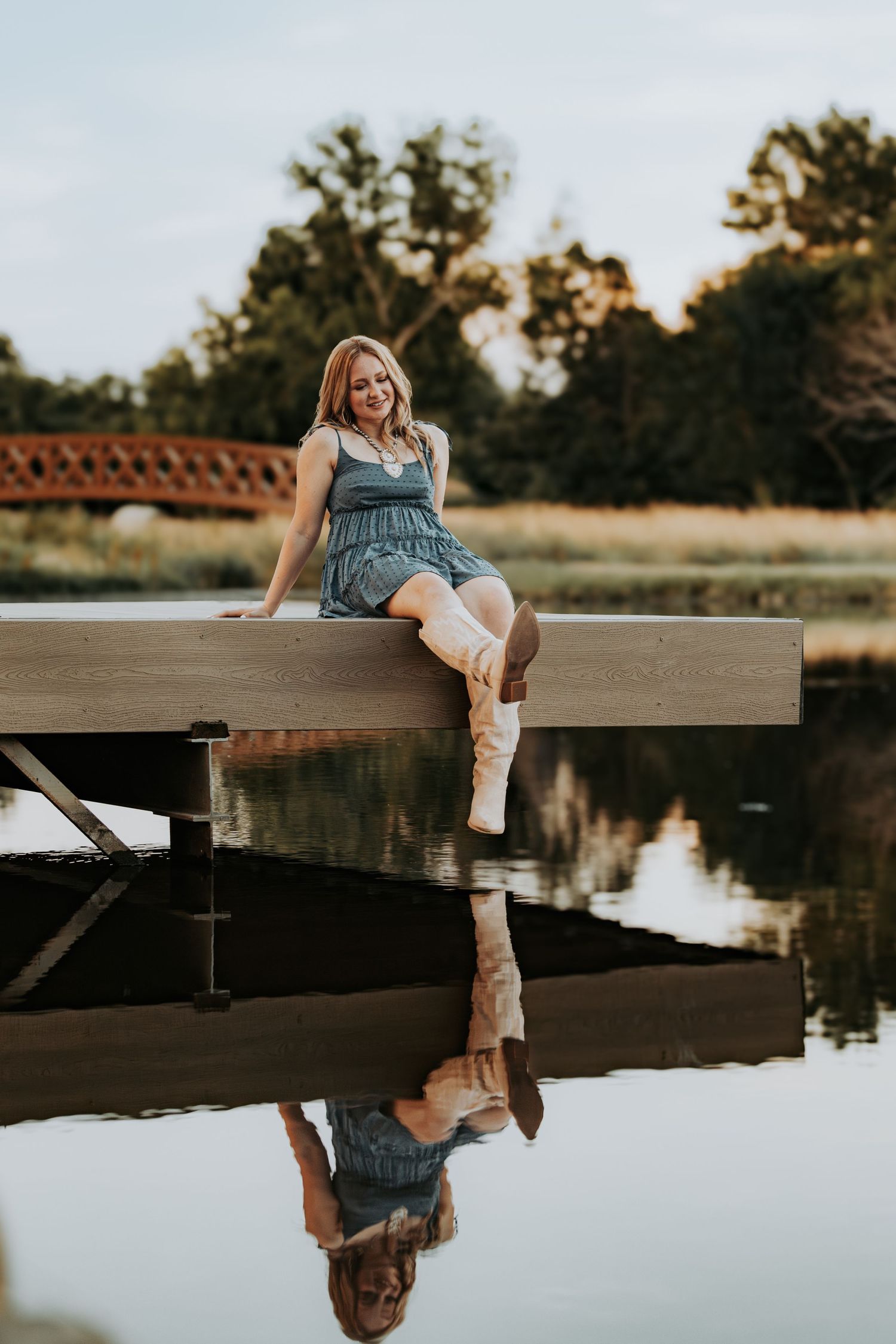 A person sits on a wooden dock by a tranquil lake at sunset while their reflection appears in the calm water below.