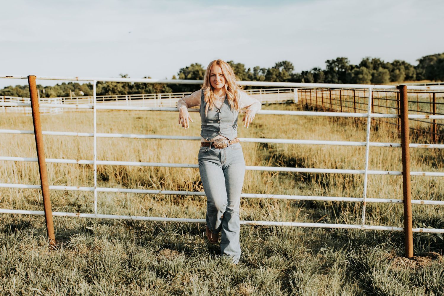Two people lean against a rustic wooden fence overlooking a golden grass field on a sunny day.