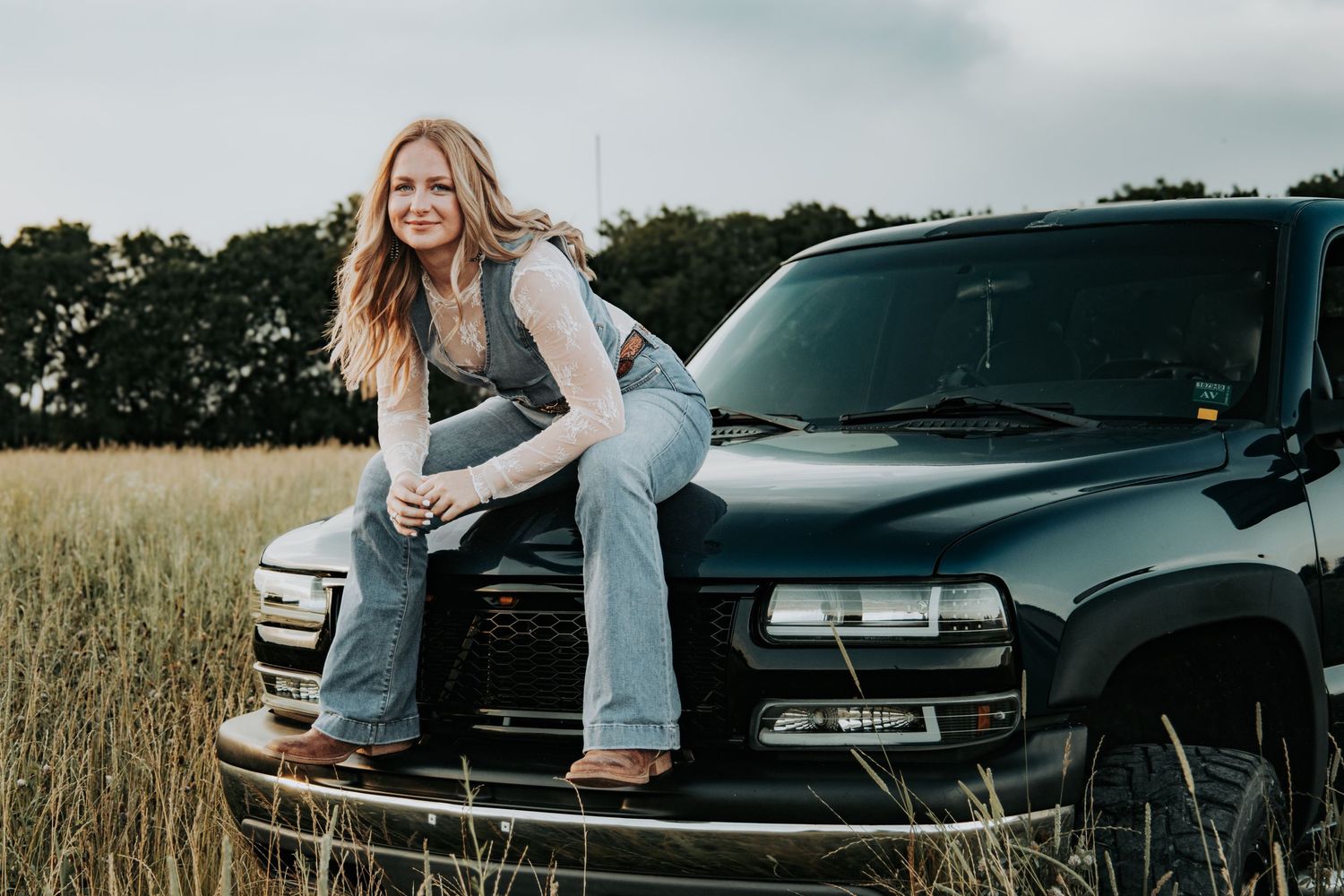 A couple embraces during a romantic photoshoot near a black car in a golden wheat field at dusk.