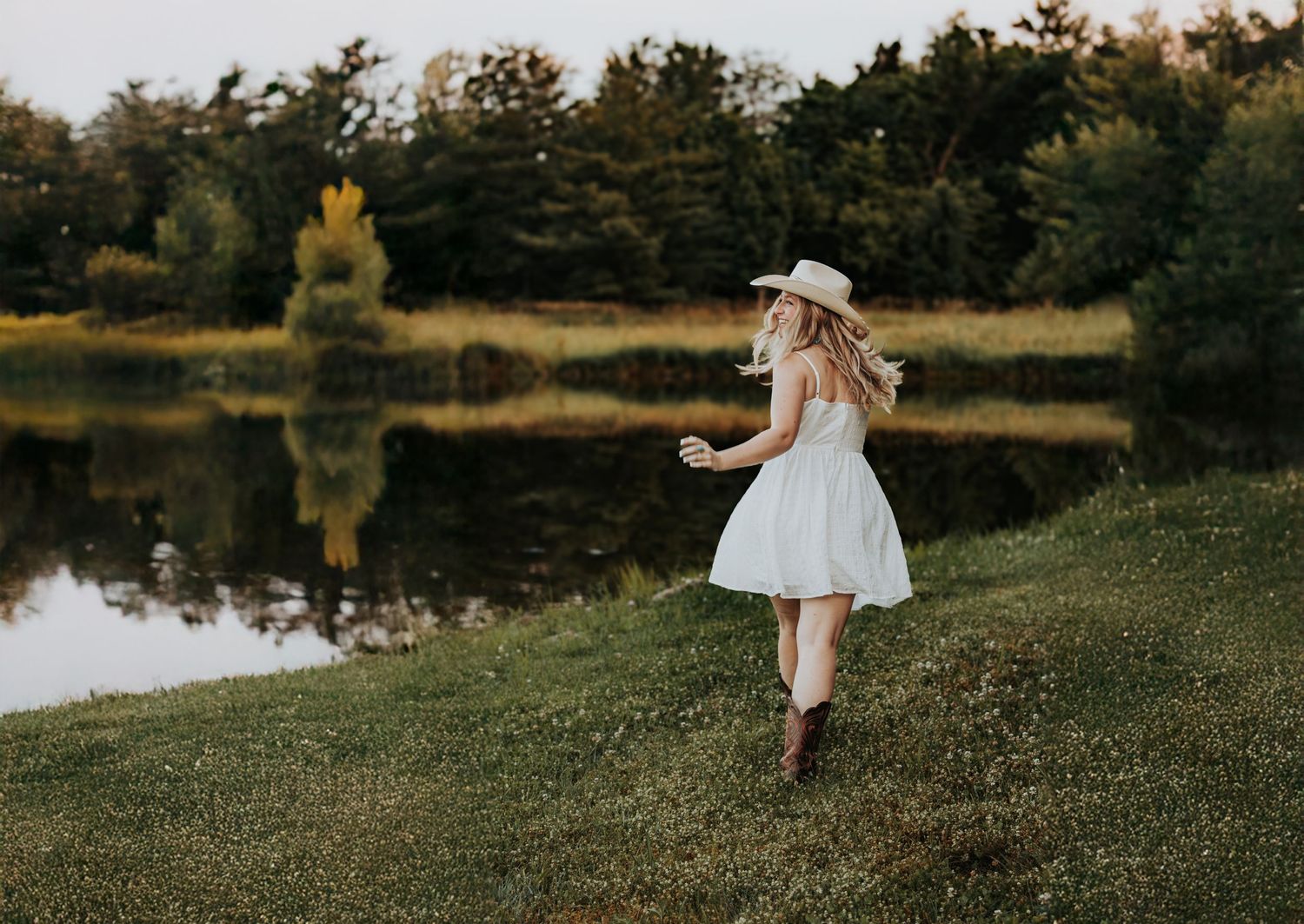 A child wearing a white dress runs playfully through a grassy field near a scenic pond at sunset.